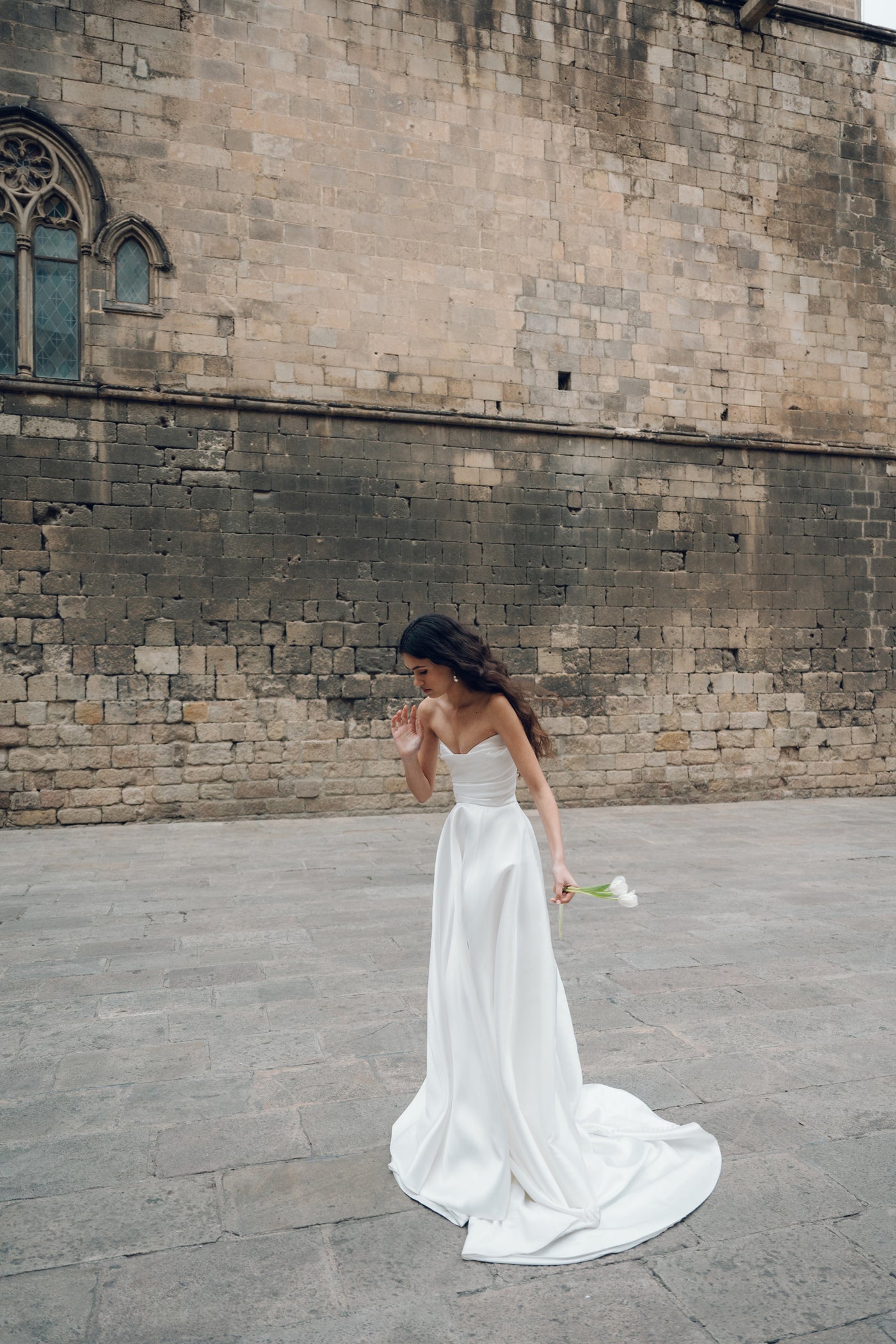 A woman in a strapless Marisol - Jenny Yoo Wedding Dress from Bergamot Bridal, featuring a ball skirt, holds a single white flower as she walks on a stone pavement in front of an old brick wall.