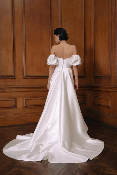 A woman in an elegant Jenny Yoo Wedding Dress gown with a draped bodice and a train, standing in a room with wood-paneled walls.