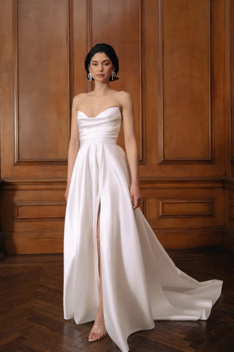 Woman in an elegant Jenny Yoo Wedding Dress with a draped bodice and a high slit, standing in a room with wooden paneling from Bergamot Bridal.