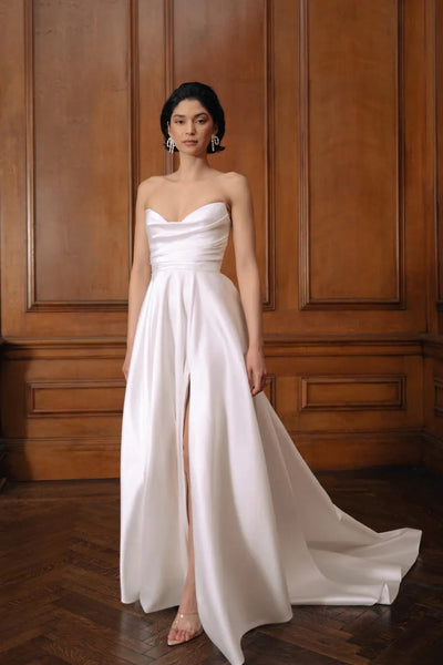 Woman in an elegant Jenny Yoo Wedding Dress with a draped bodice and a high slit, standing in a room with wooden paneling from Bergamot Bridal.