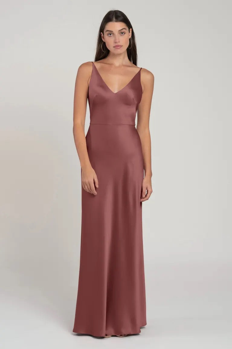 A woman models a sleeveless, V-neck, long burgundy satin Marla bridesmaid dress by Jenny Yoo against a neutral background.