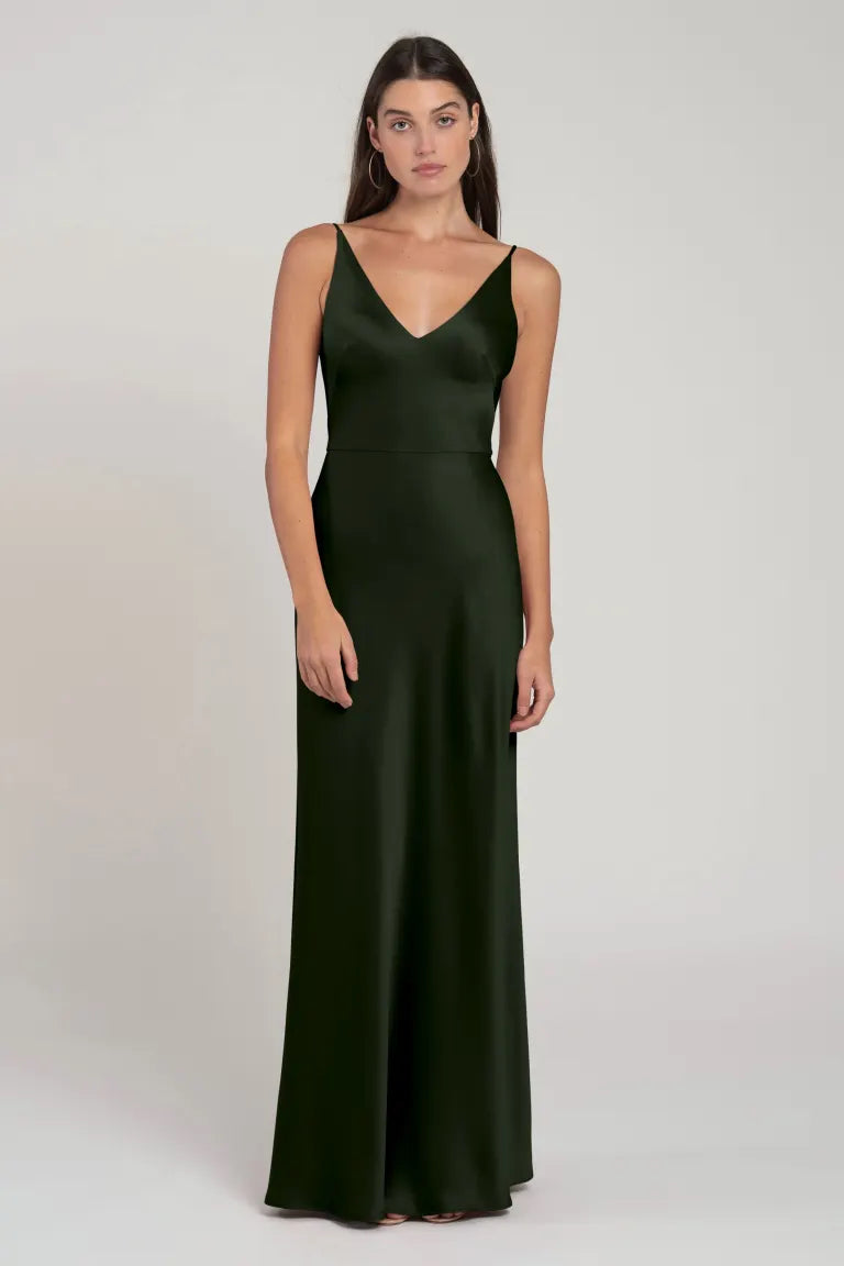 A woman stands wearing the Marla Bridesmaid Dress by Jenny Yoo from Bergamot Bridal, a long, dark green, sleeveless slip dress with a V-neckline and a natural waist seam, against a plain background.