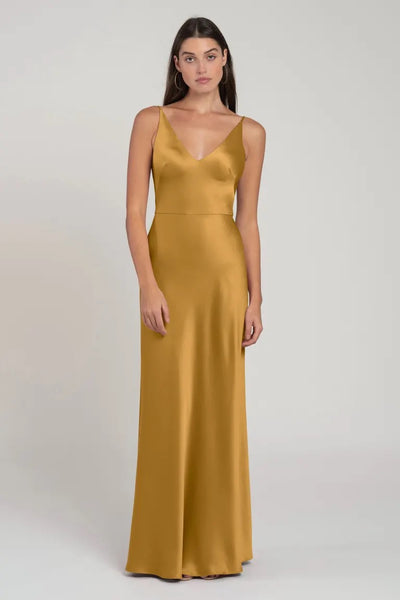 A woman modeling a long, gold satin Marla bridesmaid dress by Jenny Yoo with a bias cut skirt and v-neckline against a plain background from Bergamot Bridal.