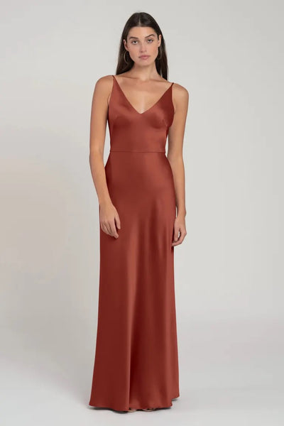 Woman posing in a plain background wearing an elegant rust-colored evening gown with spaghetti straps and a flattering V-neck, the Marla Bridesmaid Dress by Jenny Yoo from Bergamot Bridal.