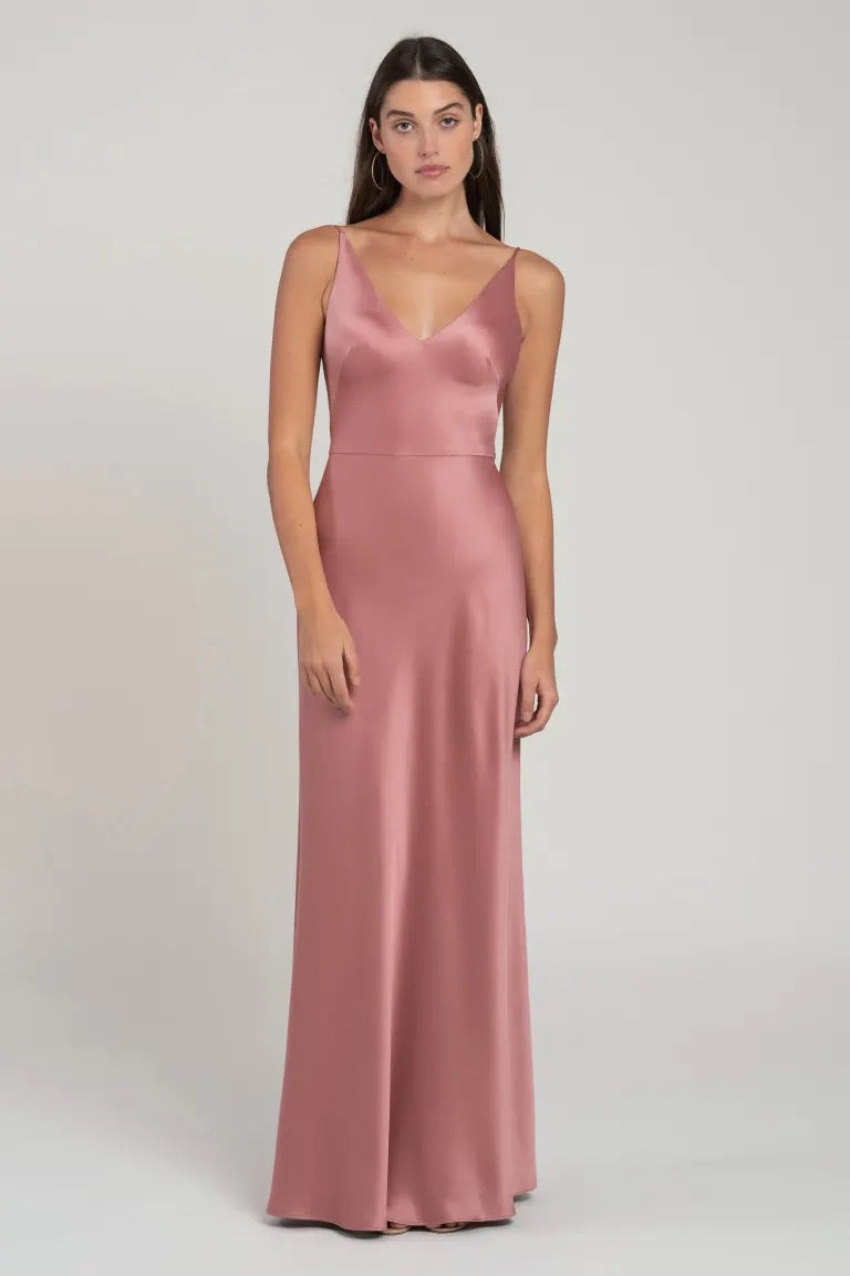 A woman posing in a plain background wearing a Marla bridesmaid dress by Jenny Yoo in pink satin with a bias-cut skirt and a V-neckline from Bergamot Bridal.