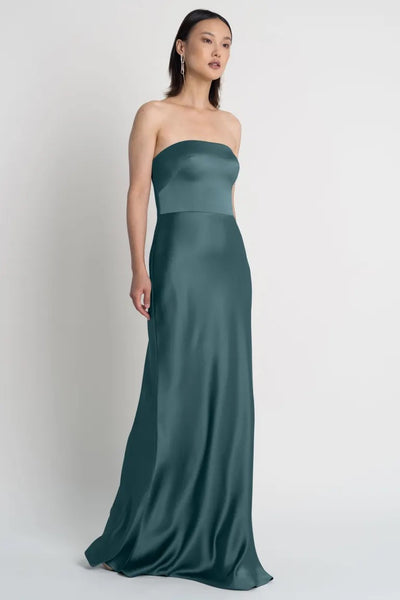 A woman in a strapless Melody bridesmaid dress by Jenny Yoo with a bias-cut skirt stands against a plain background from Bergamot Bridal.