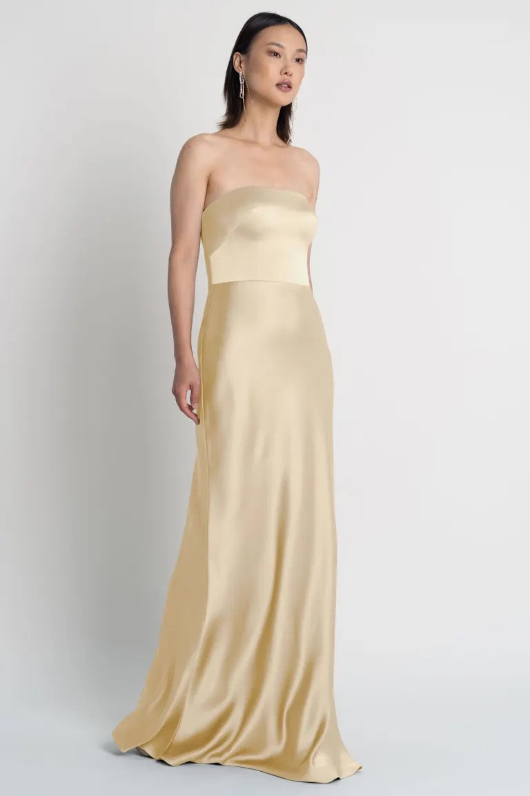 Woman in a strapless satin gold evening gown with a bias cut skirt, standing against a plain background. 
Product Name: Melody - Bridesmaid Dress by Jenny Yoo
Brand Name: Bergamot Bridal