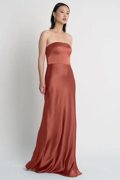 Woman in a strapless terracotta Melody bridesmaid dress by Jenny Yoo, standing against a neutral background from Bergamot Bridal.