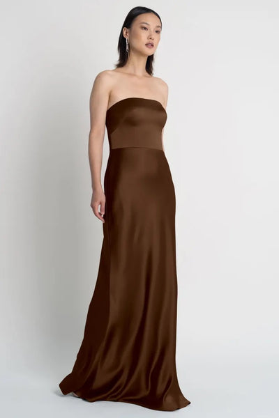 Woman in an elegant, strapless neckline, brown Melody - Bridesmaid Dress by Jenny Yoo gown standing against a neutral background.
