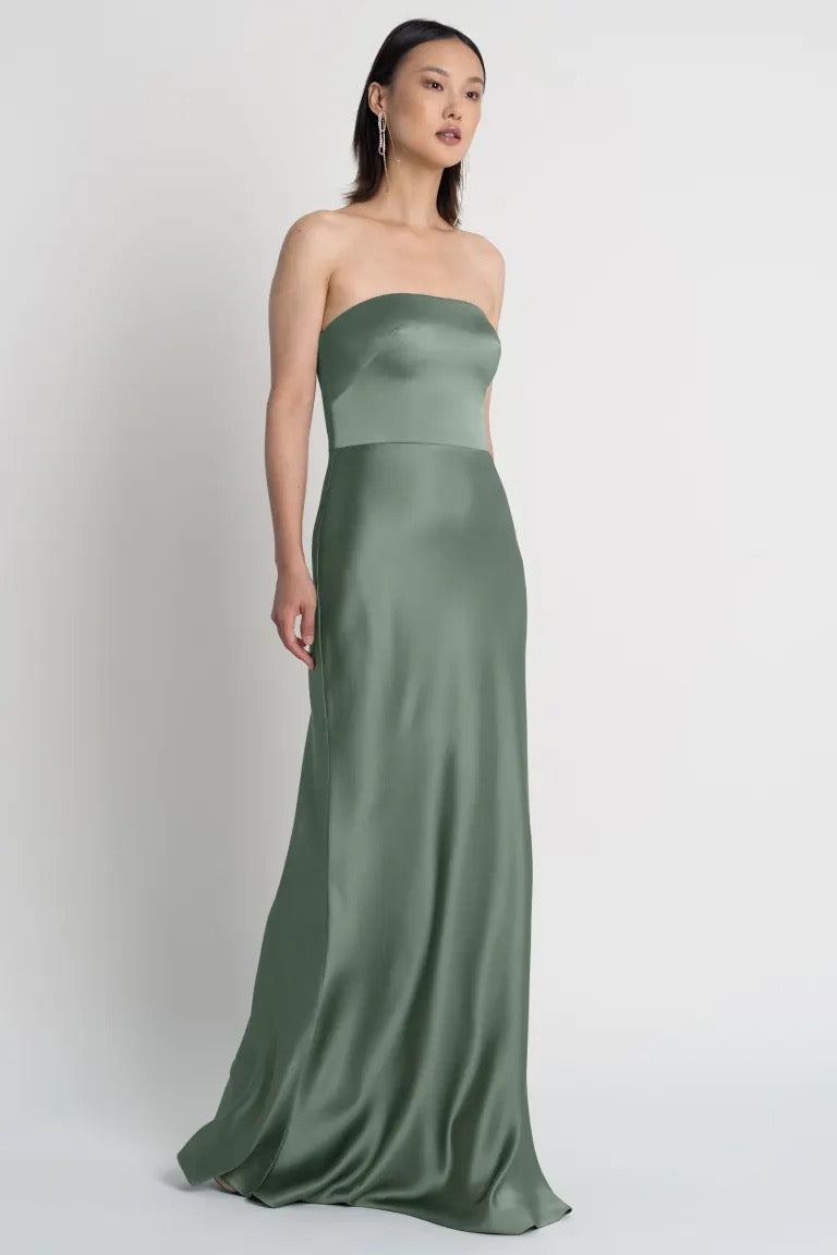 A woman in an elegant strapless Melody - Bridesmaid Dress by Jenny Yoo with a bias cut skirt stands against a plain background from Bergamot Bridal.