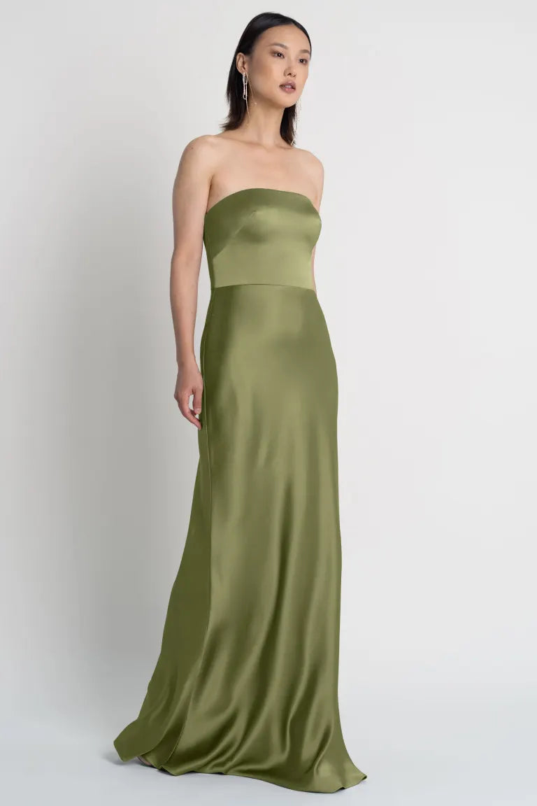 A woman stands against a plain background wearing the Melody - Bridesmaid Dress by Jenny Yoo from Bergamot Bridal, a sleeveless, strapless, green satin dress with a bias cut skirt.