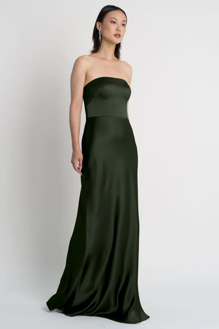 A woman stands against a plain background, wearing the Melody Bridesmaid Dress by Jenny Yoo from Bergamot Bridal, featuring a strapless neckline and a dark green bias-cut satin skirt.