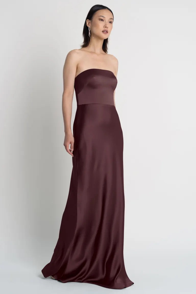 Woman in a strapless neckline, maroon Melody - Bridesmaid Dress by Jenny Yoo satin dress standing against a gray background.