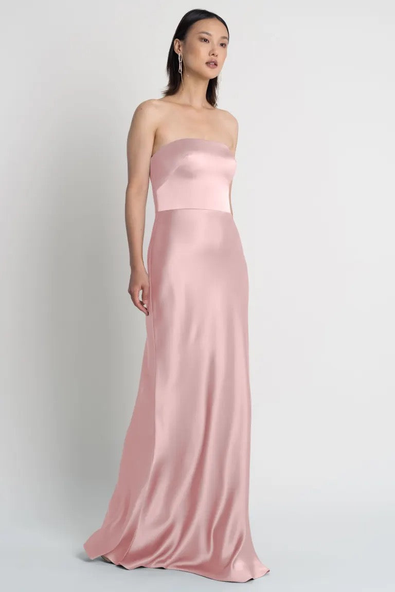 Woman in a strapless blush Melody satin dress by Jenny Yoo with a bias-cut skirt standing against a plain background from Bergamot Bridal.