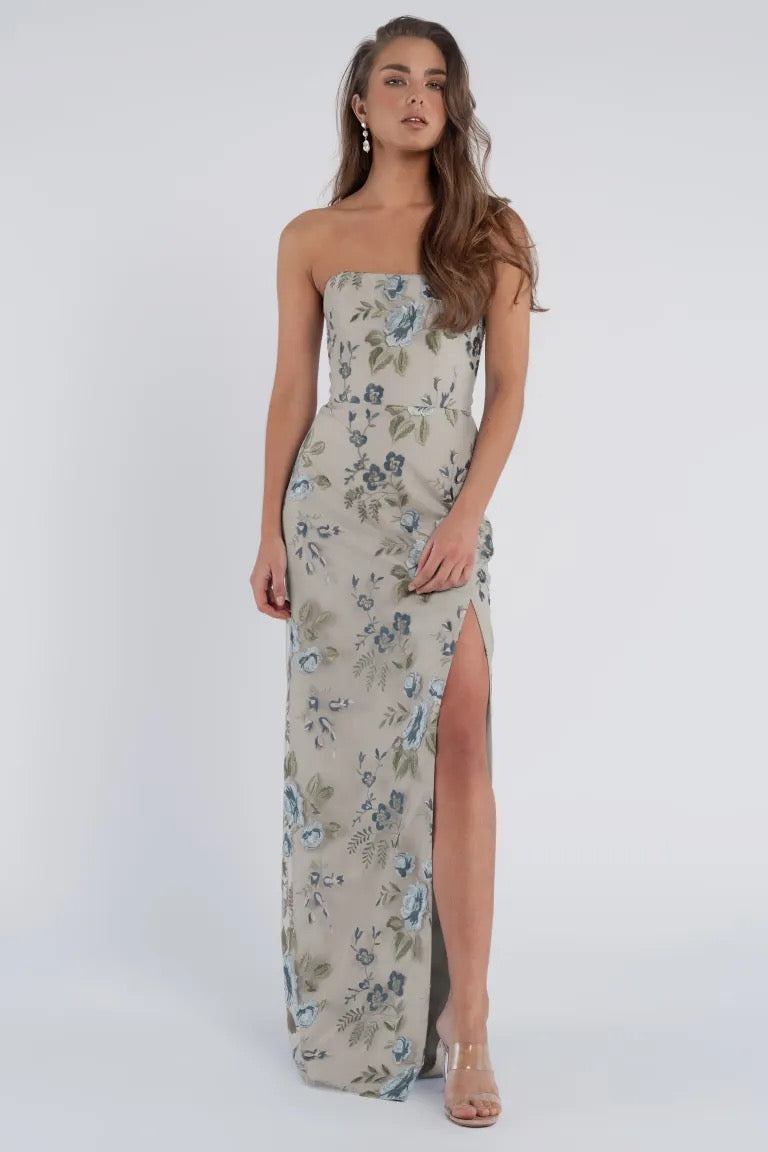 A woman is seen standing in front of a white background, donning a strapless Mercy Bridesmaid Dress by Jenny Yoo from Bergamot Bridal. The floral-print dress features a modern straight neckline and a stylish side slit, complemented perfectly with clear heels.