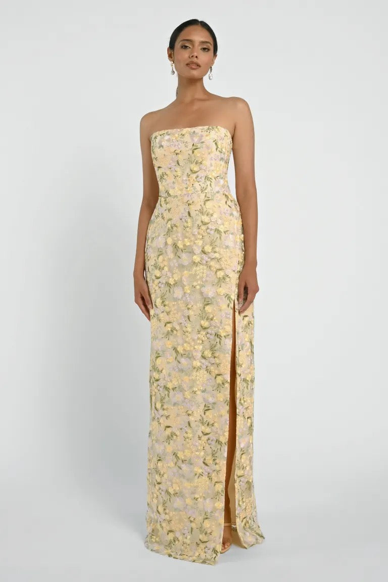 A person is wearing a strapless Mercy Bridesmaid Dress by Jenny Yoo from Bergamot Bridal, featuring a floral pattern and a high slit. The modern straight neckline accentuates the design as they stand against a plain background.