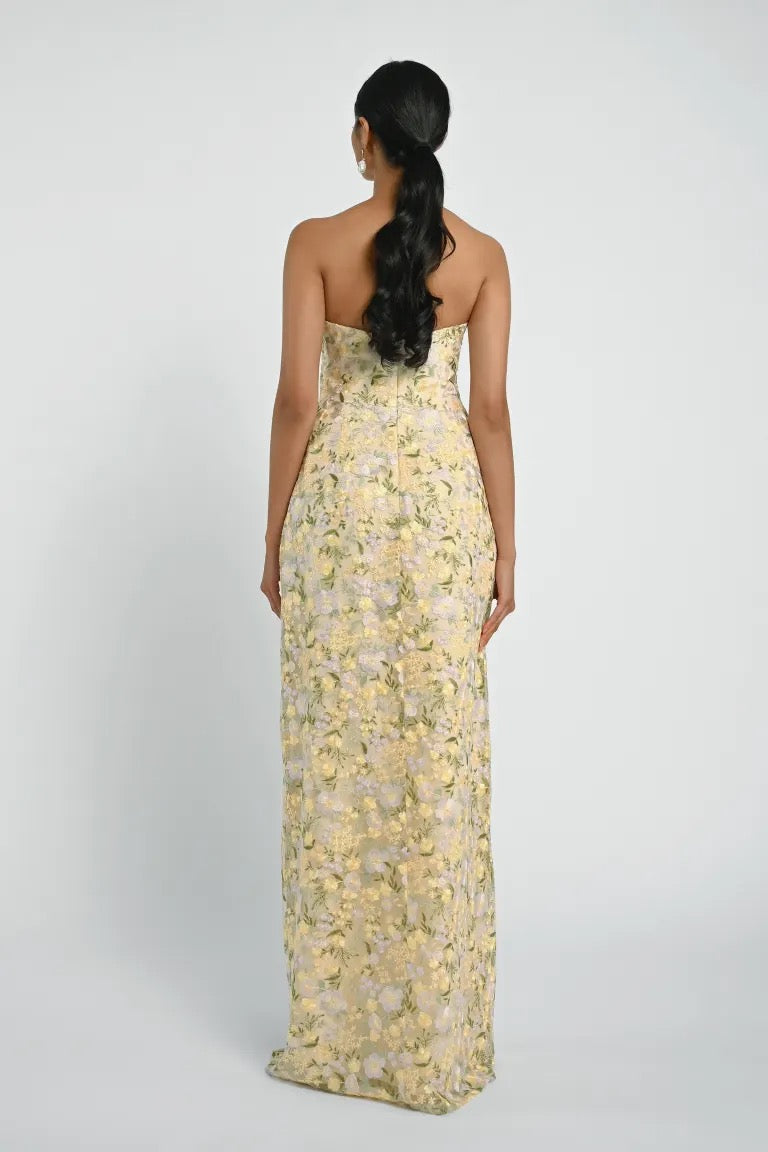 A woman with long, dark hair in a ponytail is facing away from the camera, wearing the Mercy Bridesmaid Dress by Jenny Yoo from Bergamot Bridal, which features a strapless, floor-length silhouette with a floral pattern and an embroidered tulle overlay.