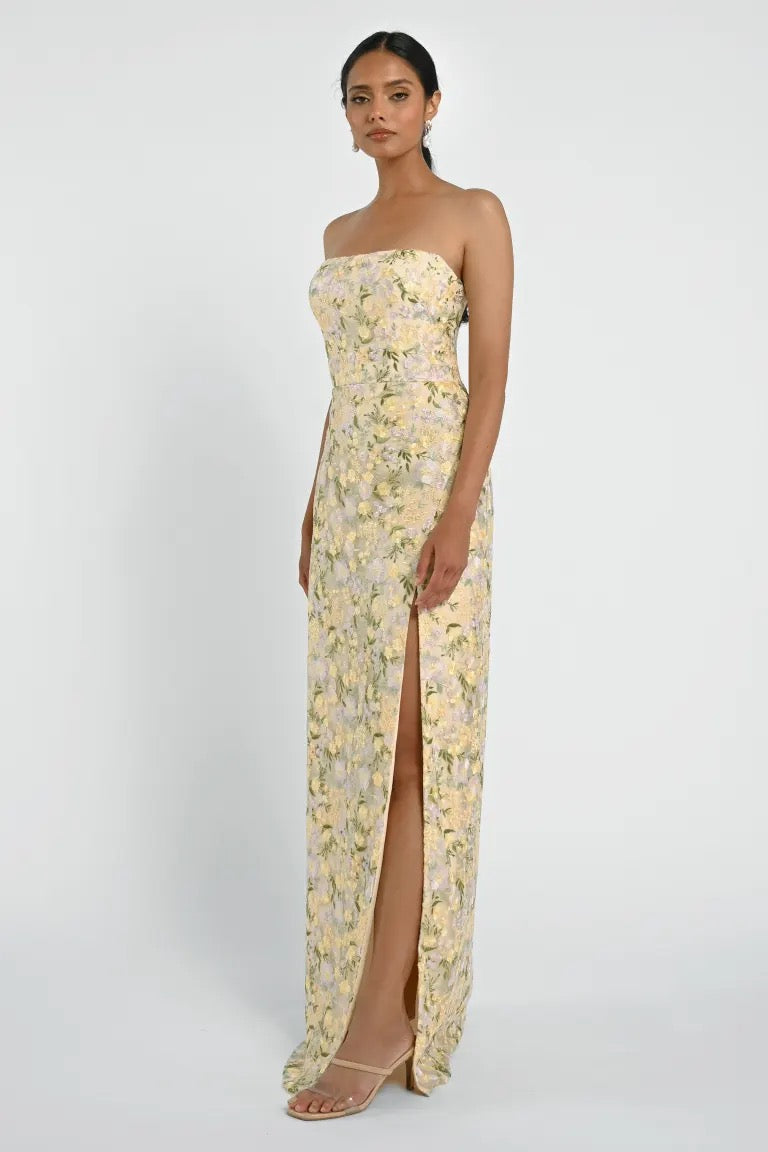 A person stands in a floral strapless Mercy Bridesmaid Dress by Jenny Yoo, from Bergamot Bridal, with a thigh-high slit and modern straight neckline, looking forward. The background is plain white.