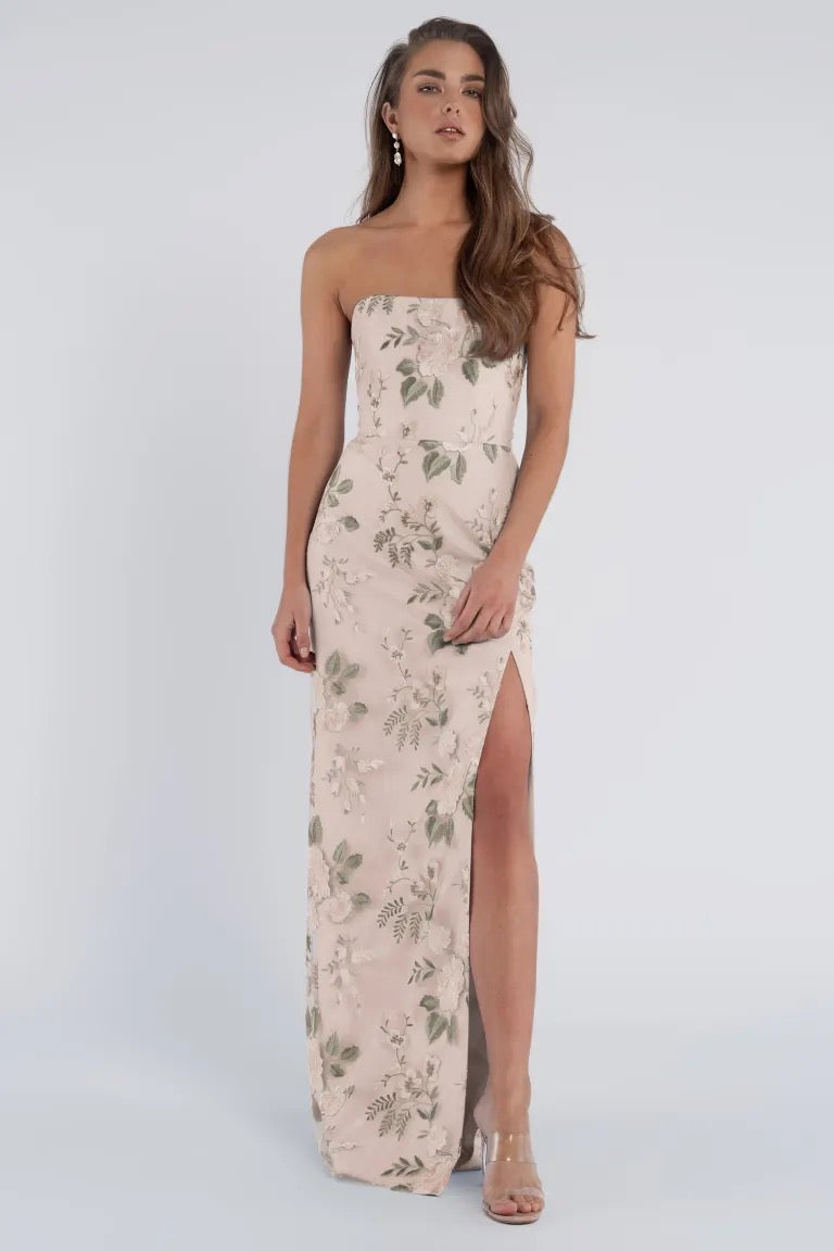 A woman is wearing the strapless, floor-length Mercy Bridesmaid Dress by Jenny Yoo from Bergamot Bridal. It features a modern straight neckline and a high slit on the right side. The floral design enhances the elegance of its embroidered tulle fabric. She has long hair and is standing against a plain background.