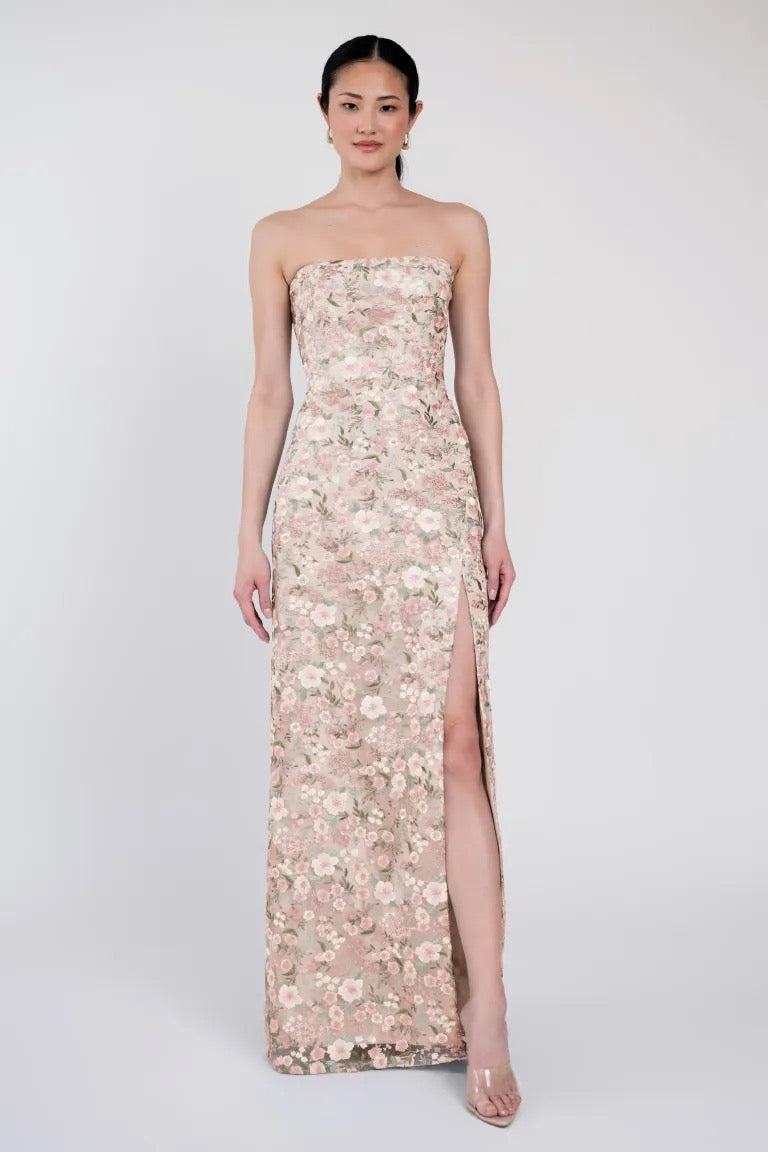 A woman in the sophisticated Mercy Bridesmaid Dress by Jenny Yoo from Bergamot Bridal, showcasing a floor-length floral pattern with a high slit and a contemporary straight neckline, stands against a plain background.