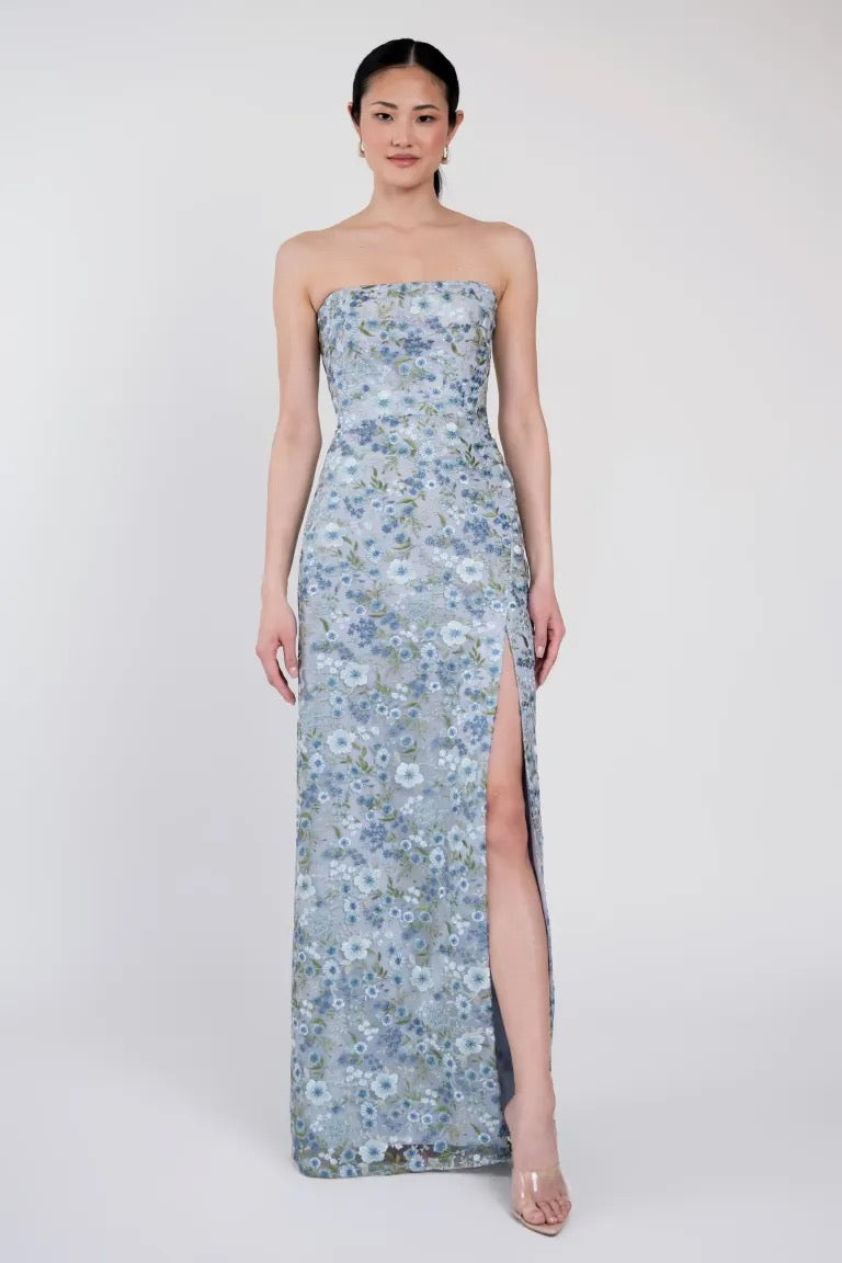 A woman stands wearing the Mercy Bridesmaid Dress by Jenny Yoo from Bergamot Bridal, a strapless, floor-length gown with a blue floral pattern, featuring a thigh-high slit and modern straight neckline. She is in a well-lit, plain setting, facing forward.