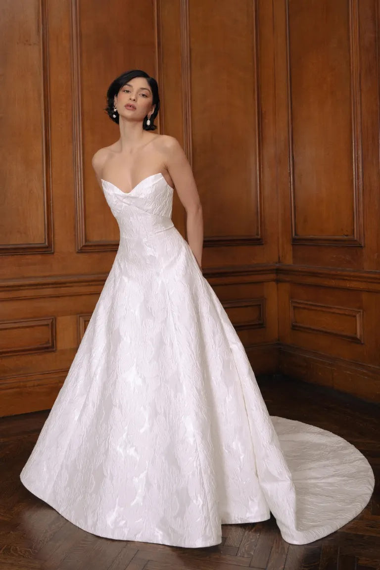 Woman posing in an elegant Bergamot Bridal Jenny Yoo Wedding Dress featuring off-the-shoulder sleeves and a classic wooden panel background.