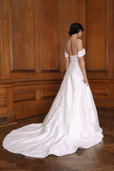 Bride in an elegant white Bergamot Bridal Jenny Yoo Wedding Dress made of floral jacquard fabric with off-the-shoulder sleeves, standing in a wood-paneled room.