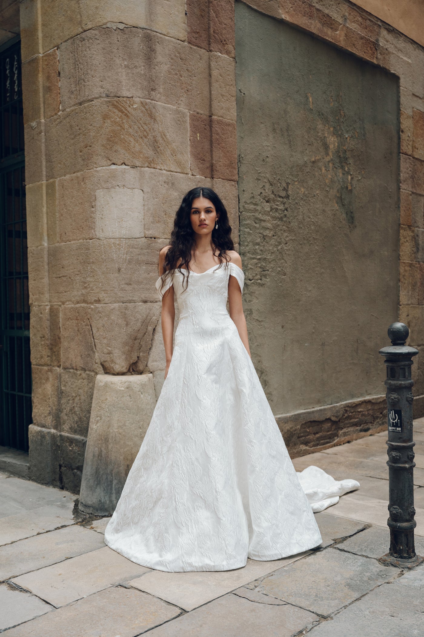 Wearing the Michele - Jenny Yoo Wedding Dress by Bergamot Bridal, which features a white, off-shoulder design with detachable sleeves, a woman stands by a stone building on a cobblestone street.