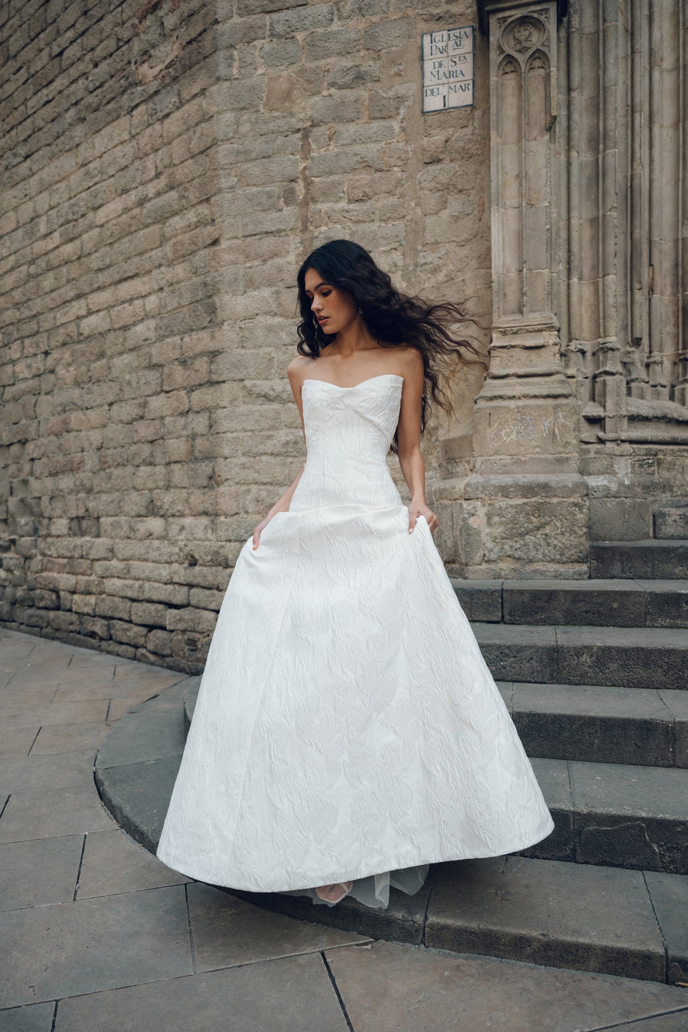 A woman in the Michele - Jenny Yoo Wedding Dress by Bergamot Bridal, a strapless white gown made from floral jacquard fabric, stands on a stone path beside an old brick building, with her hair blowing in the wind.