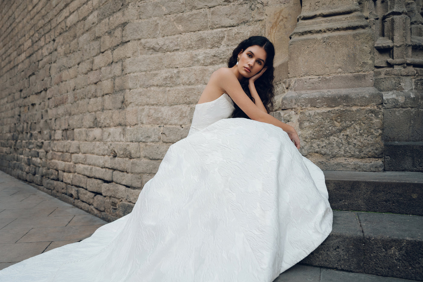 A woman wearing the Michele - Jenny Yoo Wedding Dress by Bergamot Bridal, featuring a striking silhouette and detachable off-the-shoulder sleeves, sits on stone steps beside a brick wall, gazing thoughtfully into the distance.