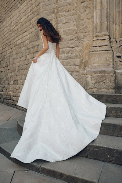 A woman in the Michele - Jenny Yoo Wedding Dress by Bergamot Bridal, featuring detachable off-the-shoulder sleeves and a long, flowing white design, stands on stone steps beside a textured stone wall, creating a striking silhouette.