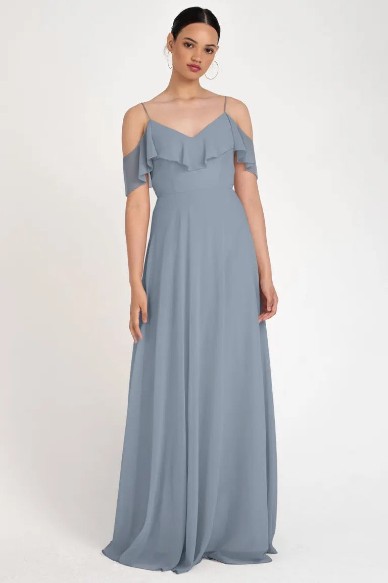 Woman in a blue off-the-shoulder neckline evening gown with a circle skirt silhouette posing against a neutral background, wearing the Mila Bridesmaid Dress by Jenny Yoo from Bergamot Bridal.