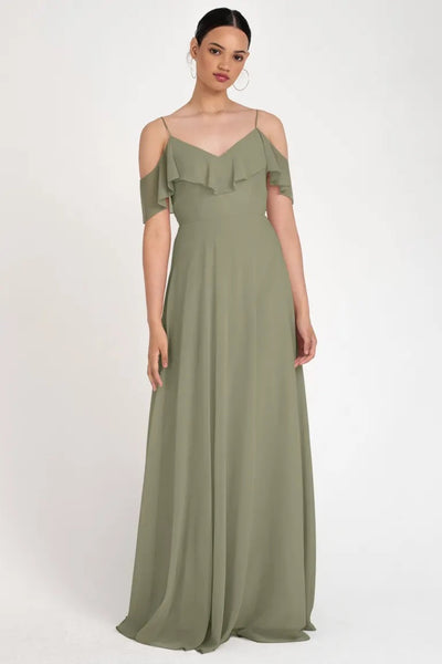 A woman in a Mila - Bridesmaid Dress by Jenny Yoo in an olive green, circle skirt silhouette gown with an off-the-shoulder neckline stands against a neutral background from Bergamot Bridal.
