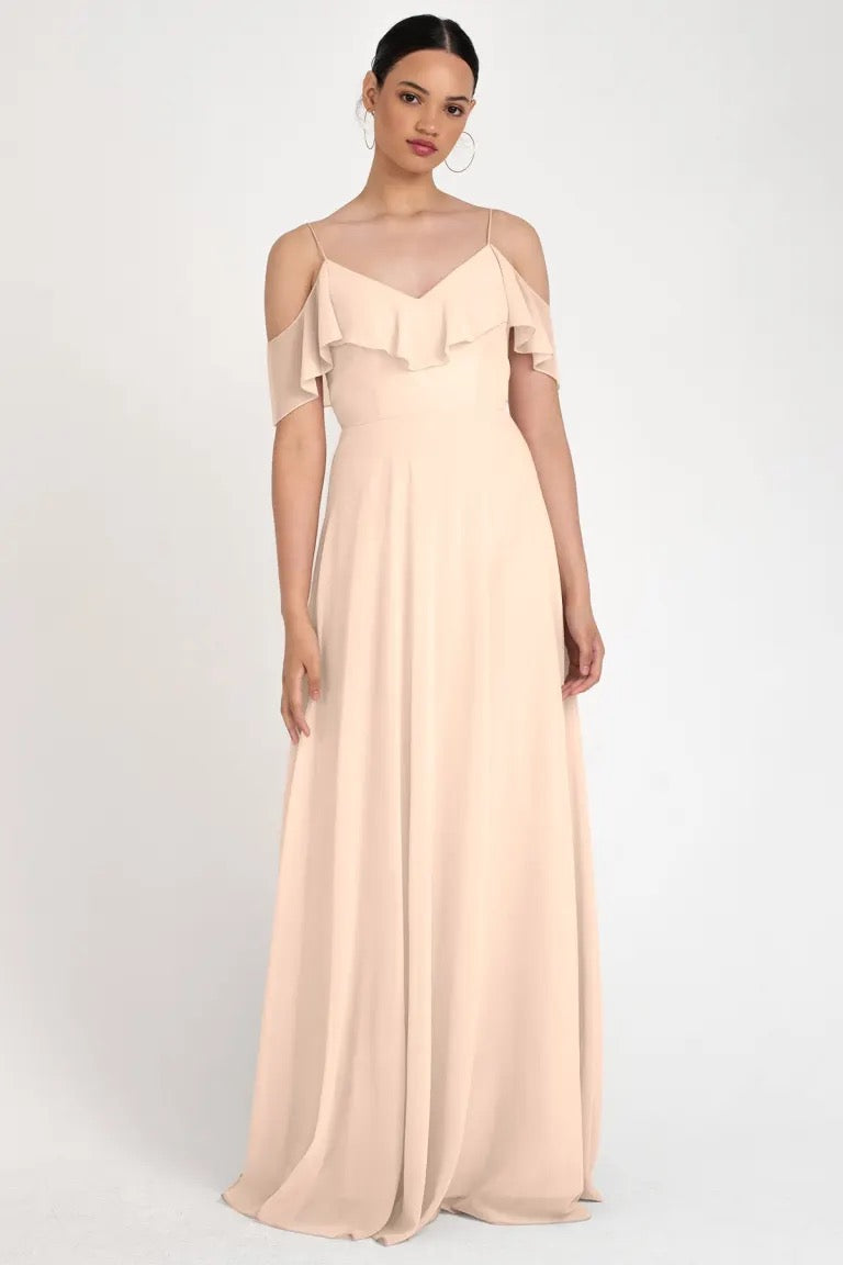 Woman in an elegant beige off-the-shoulder gown with ruffle detailing and a circle skirt silhouette, wearing the Mila Bridesmaid Dress by Jenny Yoo from Bergamot Bridal.