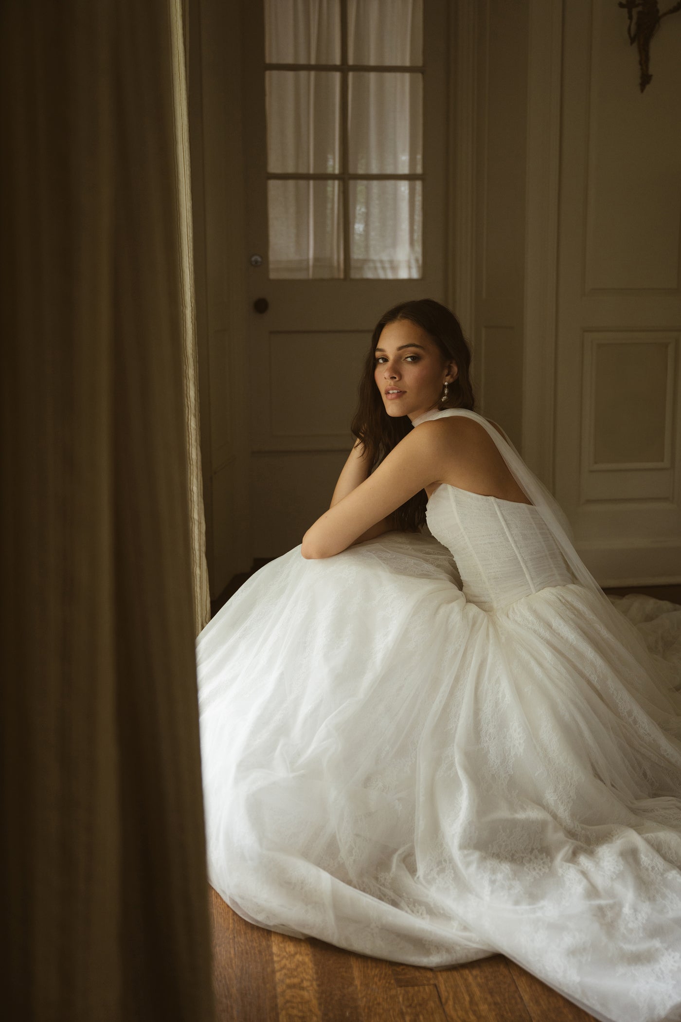 A woman in the Miriam by Jenny Yoo gown from Bergamot Bridal sits on a wooden floor near a window, looking over her shoulder in a softly lit room.