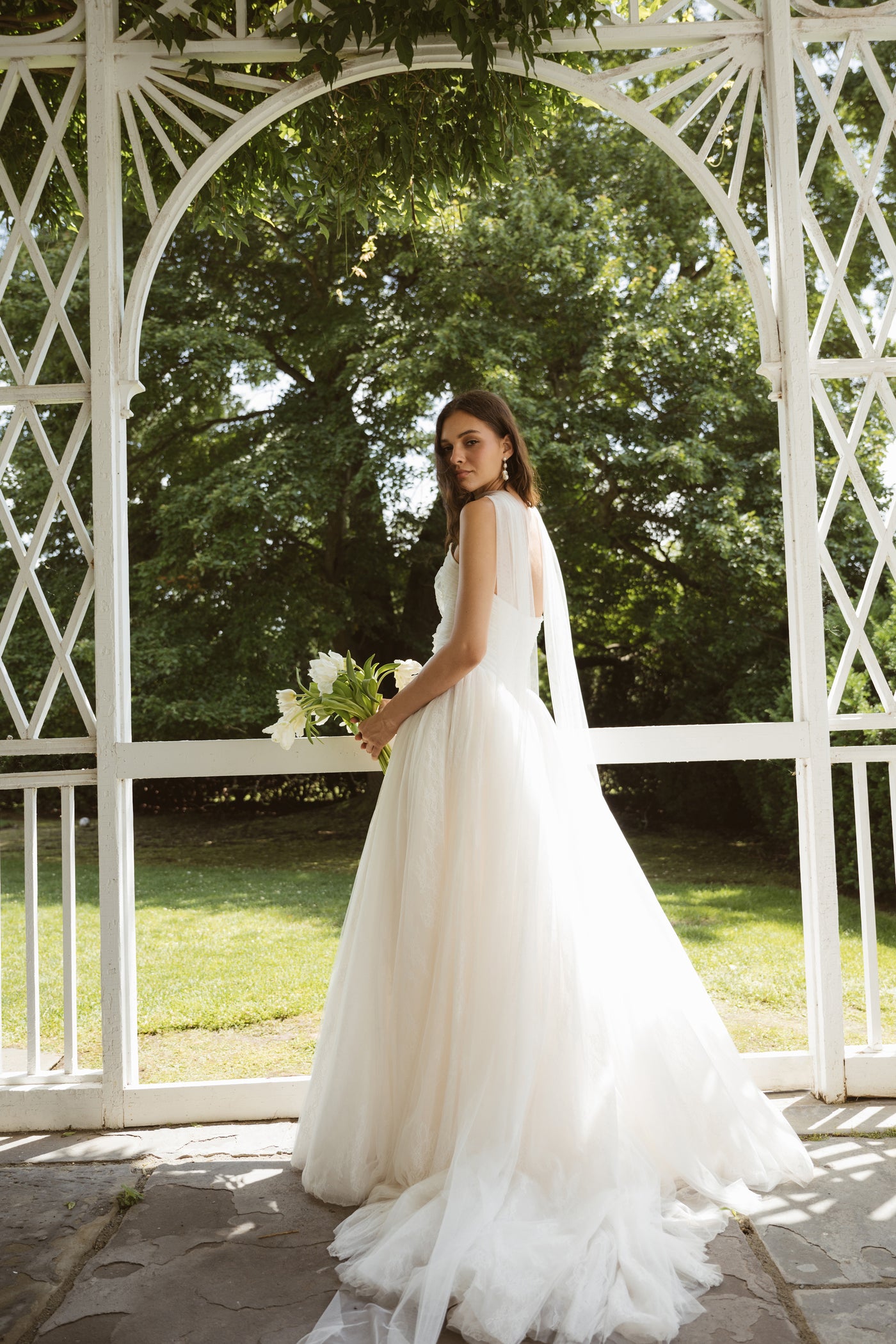 A woman wearing the Miriam by Jenny Yoo bridal gown from Bergamot Bridal stands in a gazebo, holding a bouquet amid greenery and sunlight.