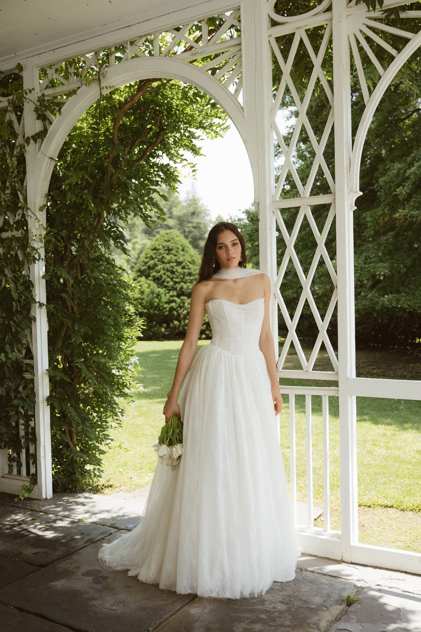A woman in the Miriam by Jenny Yoo gown from Bergamot Bridal stands under a white gazebo, holding a bouquet, surrounded by greenery and trees.