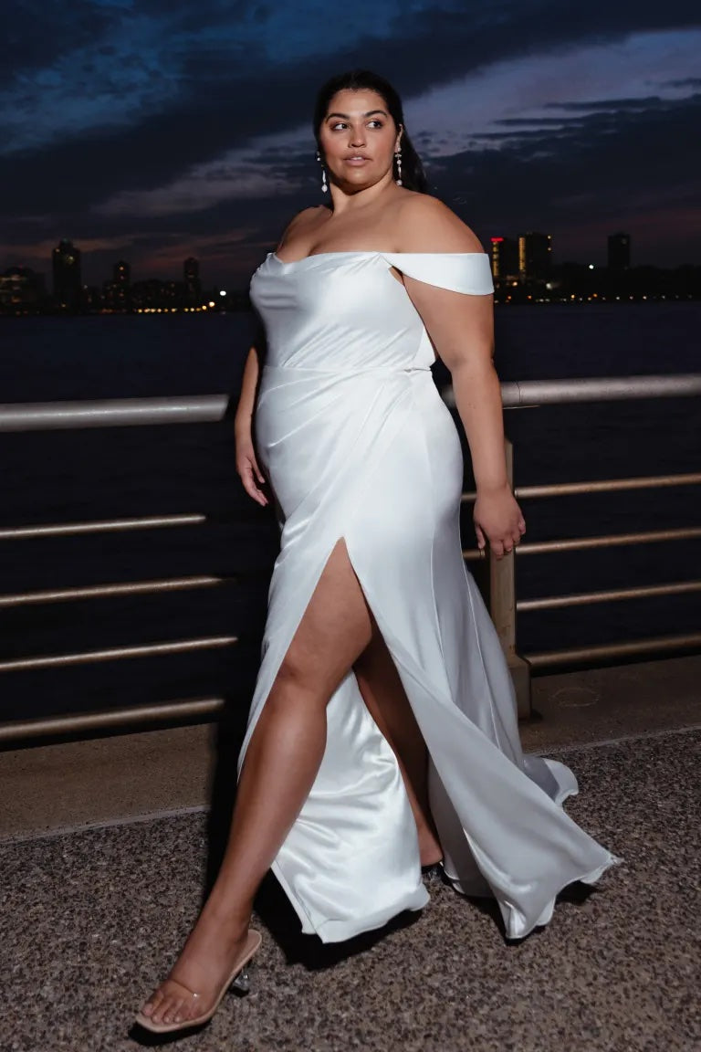A woman in an elegant Bergamot Bridal Viviana gown, characterized by its off-the-shoulder design and a thigh-high slit, stands confidently against a twilight cityscape backdrop.