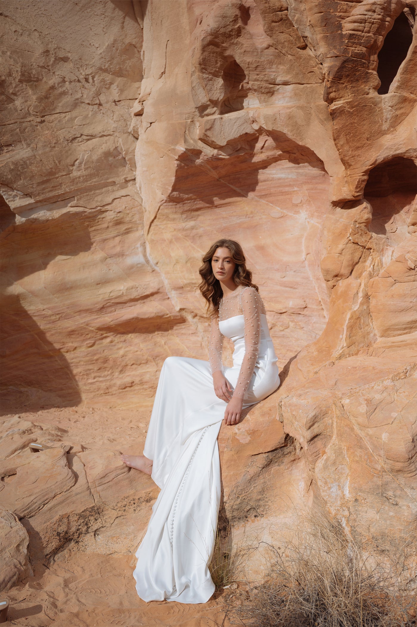 A woman in a white Jenny Yoo Wedding Dress adorned with pearls posing against a sandstone rock formation from Bergamot Bridal.