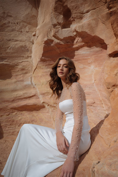 A woman in a white Jenny Yoo wedding dress with sheer sleeves and pearls sits against a sandstone rock formation from Bergamot Bridal.