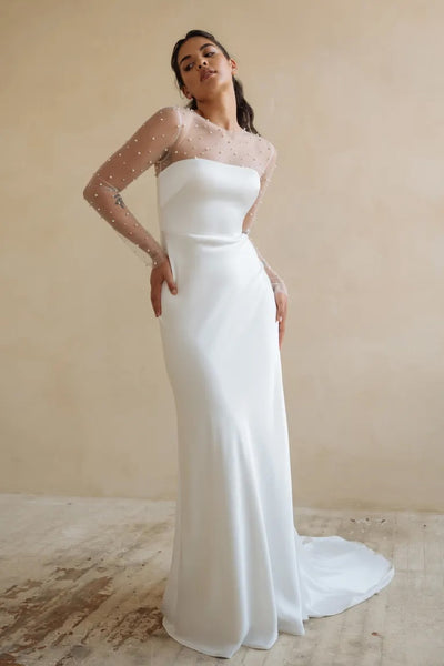 Woman in an elegant white Morgan - Jenny Yoo wedding dress with sheer dotted sleeves and pearls posing against a neutral backdrop from Bergamot Bridal.