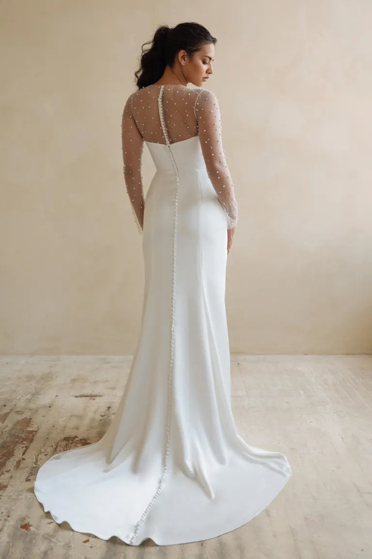 Woman in an elegant modern white Jenny Yoo wedding dress with sheer embellished sleeves adorned with pearls, standing with her back turned from Bergamot Bridal.