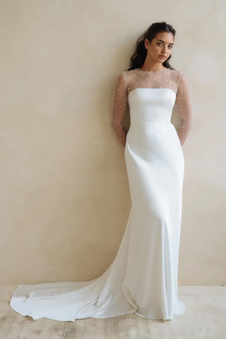 A woman in an elegant Bergamot Bridal gown with a sheer polka-dot sleeve detail and pearls as her bridal accessory, standing against a neutral backdrop.