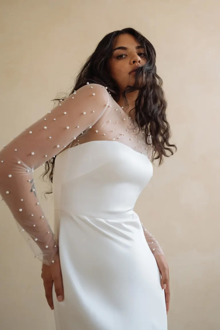 Woman in an elegant Jenny Yoo Wedding Dress by Morgan with sheer embellished sleeves and pearls posing against a neutral background.
