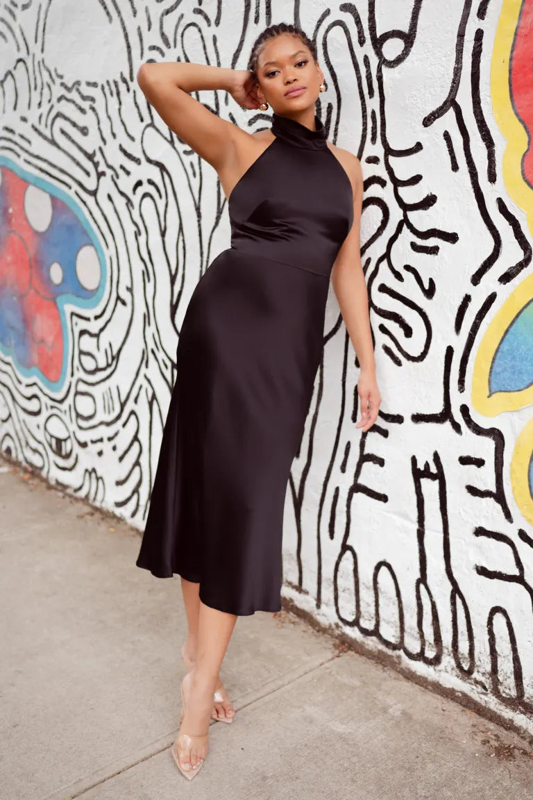 A woman in a black satin midi dress leaning against a colorful wall, showcasing a stunning Nicolette - Jenny Yoo Little White Dress from Bergamot Bridal.