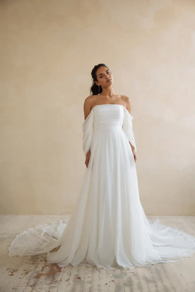 A woman in an elegant white off-shoulder Jenny Yoo Wedding Dress gown with dramatic sleeves and a long train stands against a neutral backdrop.