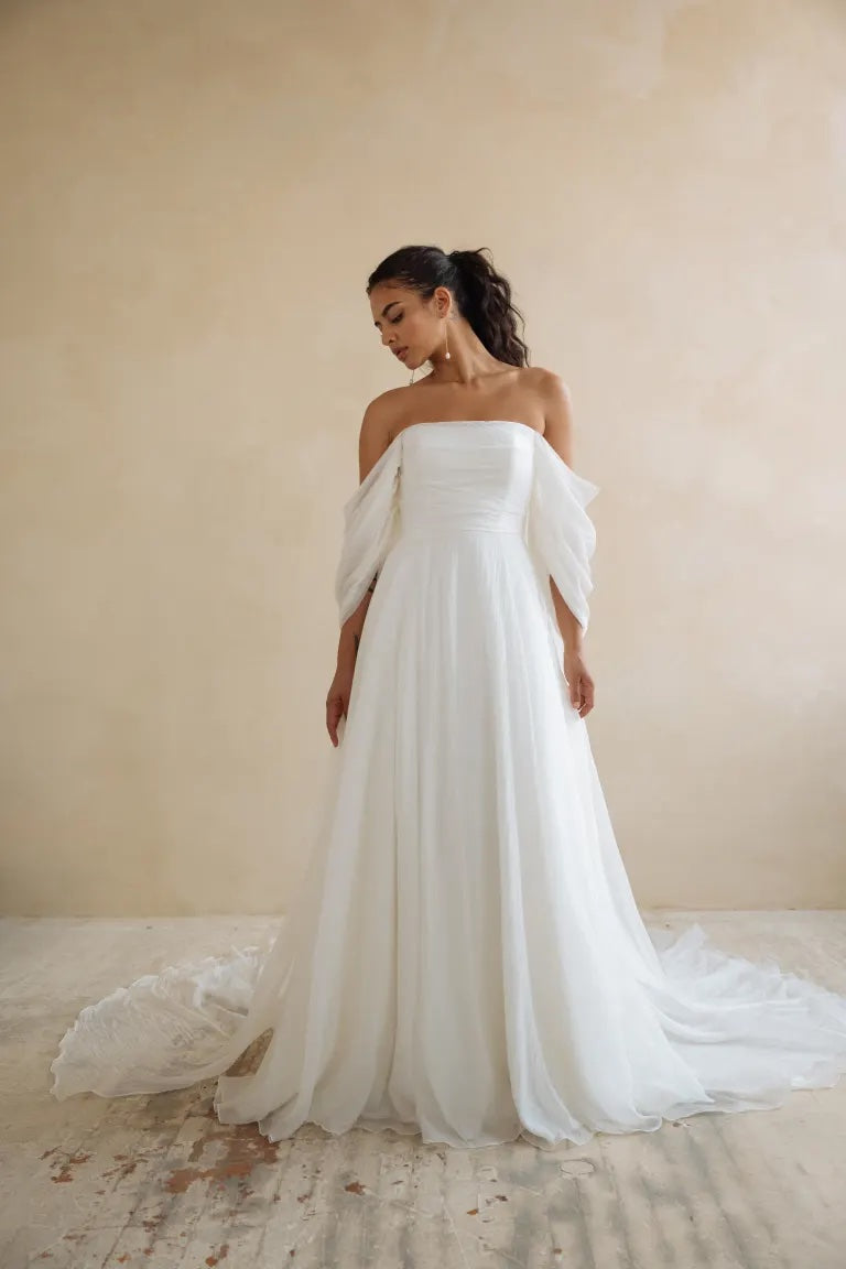 A bride in an elegant Noa - Jenny Yoo wedding dress, featuring dramatic sleeves and a flowing train, stands against a neutral backdrop.