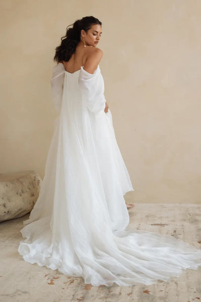 A woman in an off-the-shoulder Jenny Yoo Wedding Dress gown with dramatic sleeves and a trailing skirt, standing in a room with a neutral background from Bergamot Bridal.