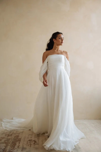 A woman in an elegant Noa - Jenny Yoo Wedding Dress gown with dramatic sleeves and a flowing train stands against a neutral backdrop.
