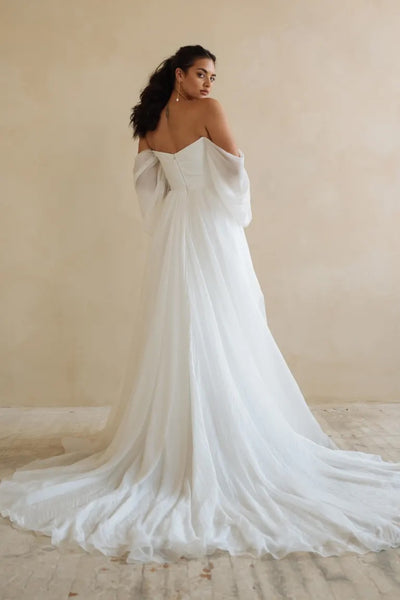 A woman in an elegant Noa - Jenny Yoo Wedding Dress gown from Bergamot Bridal, featuring dramatic sleeves, looks over her shoulder.
